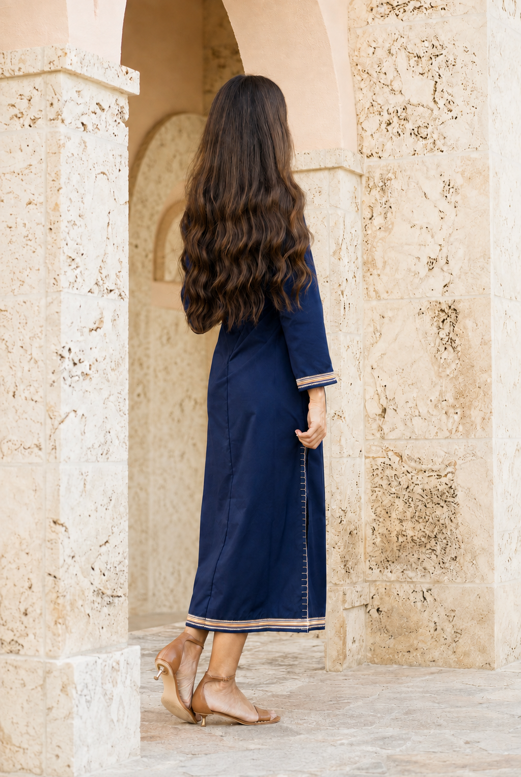 A woman with long, wavy dark brown hair is seen from behind, wearing a navy blue maxi dress with beige trim, standing in a stone archway.
