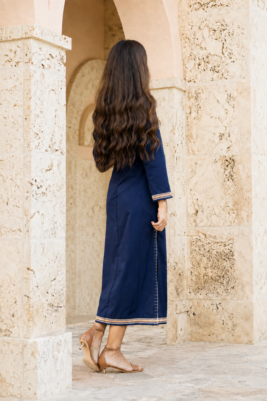 A woman with long, wavy dark brown hair is seen from behind, wearing a navy blue maxi dress with beige trim, standing in a stone archway.