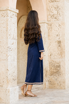 A woman with long, wavy dark brown hair is seen from behind, wearing a navy blue maxi dress with beige trim, standing in a stone archway.