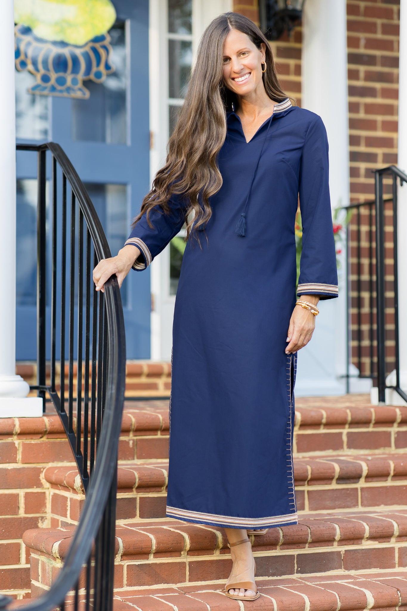 A woman wearing a navy blue kaftan dress with tan and white embroidery along the hem, cuffs and neckline. 