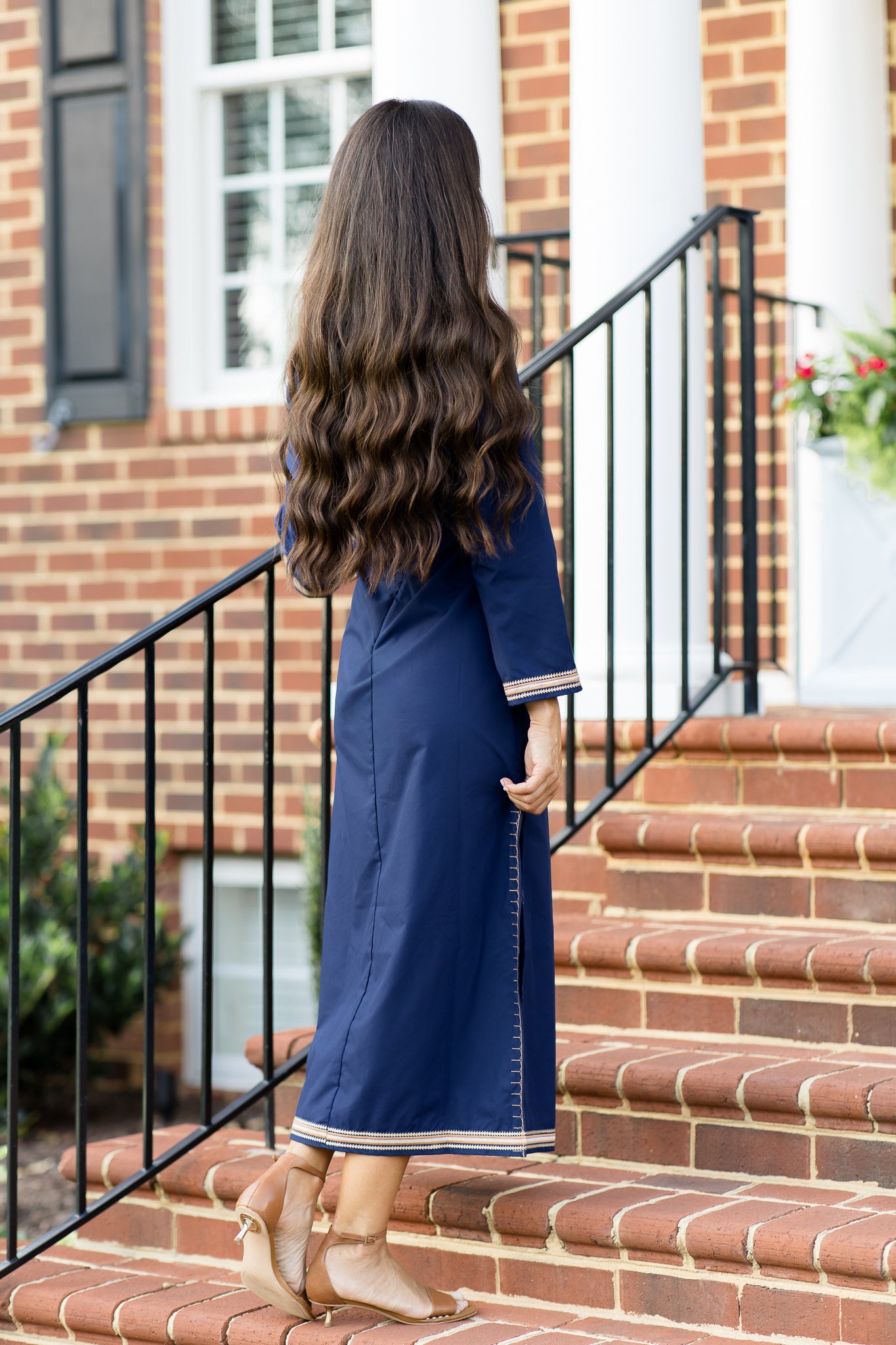 Back view of a woman wearing a navy blue kaftan dress with tan and white embroidery along the hem, cuffs and neckline. 